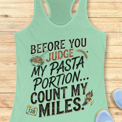 A mint racerback tank top featuring  the text 'before you judge my pasta portion...count my miles ' printed on the front, displayed flat on a light wood background.