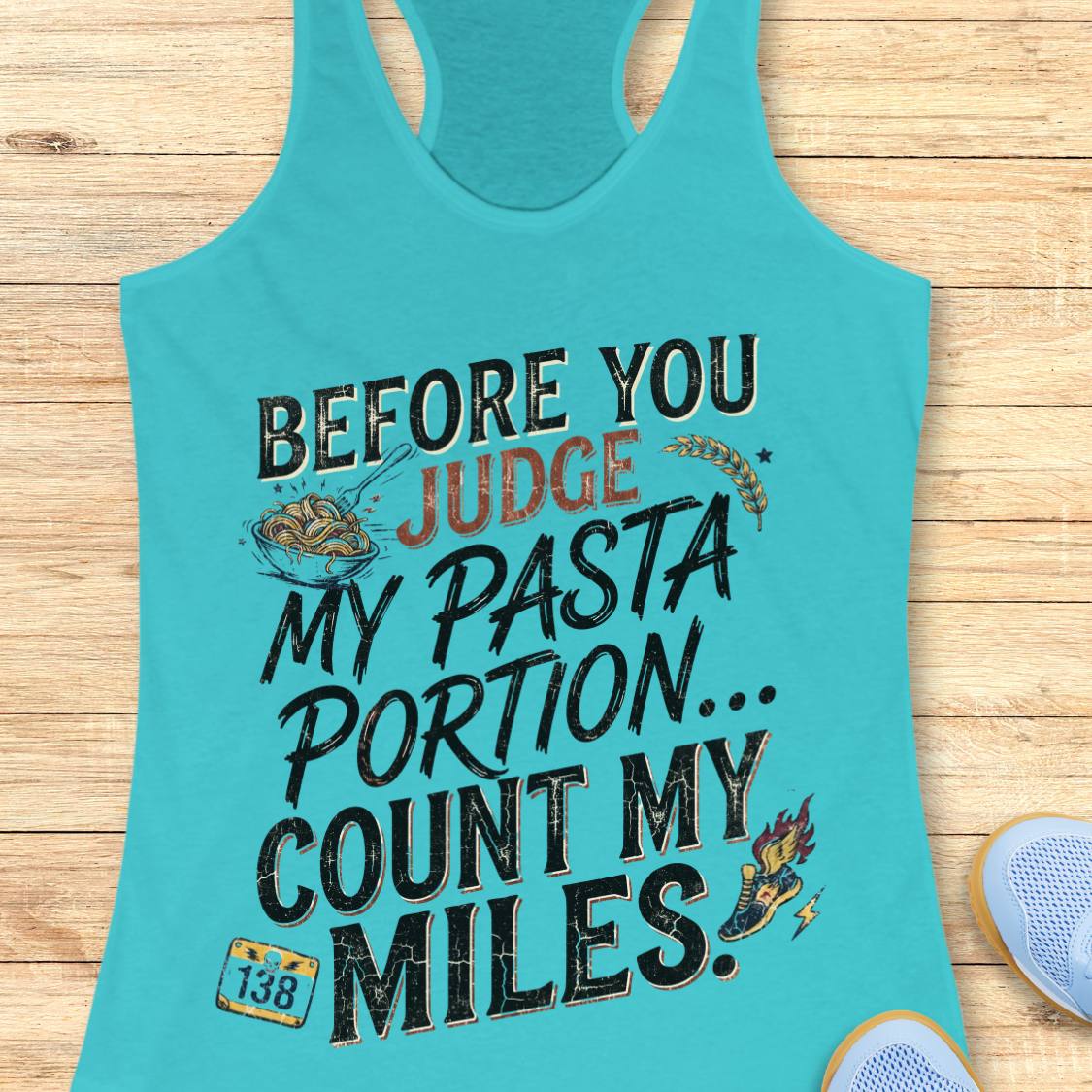 A blue racerback tank top featuring  the text 'before you judge my pasta portion...count my miles ' printed on the front, displayed flat on a light wood background.