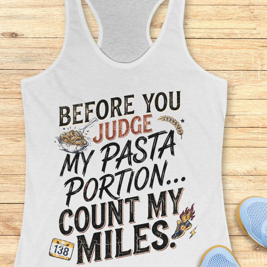 A white racerback tank top featuring  the text 'before you judge my pasta portion...count my miles ' printed on the front, displayed flat on a light wood background.