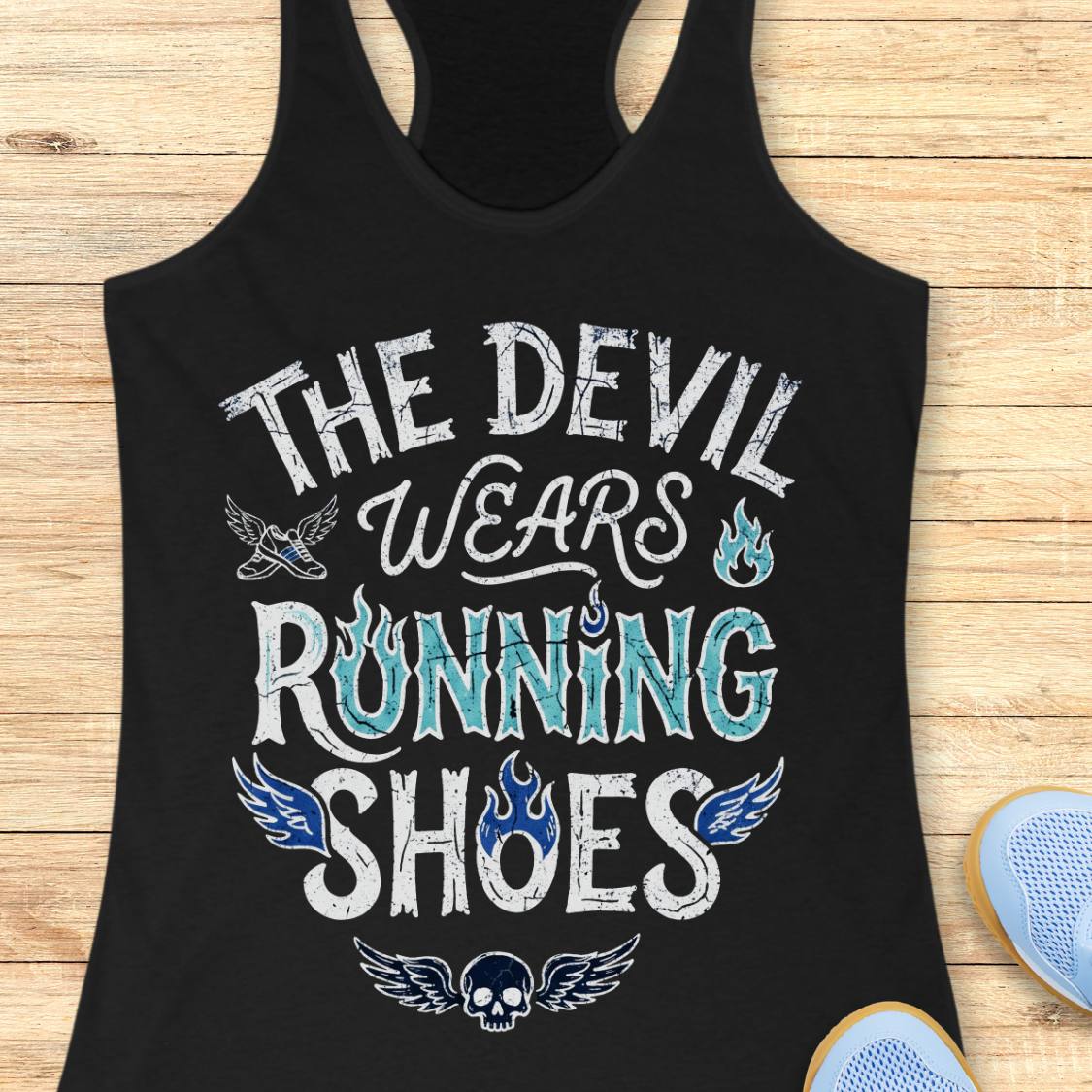 A black racerback tank top laid flat on a light wood background, featuring the graphic text 'THE DEVIL WEARS RUNNING SHOES' in distressed black and red lettering with small flame and wing illustrations. Partial blue shoes are visible at the bottom right as styling props.