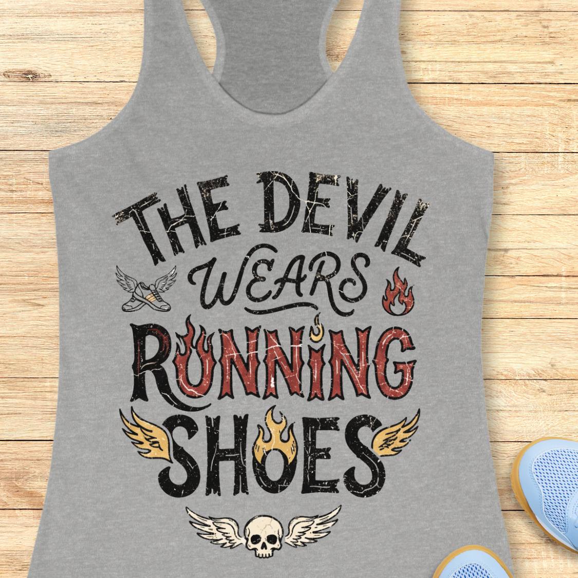 A heather gray racerback tank top laid flat on a light wood background, featuring the graphic text 'THE DEVIL WEARS RUNNING SHOES' in distressed black and red lettering with small flame and wing illustrations. Partial blue shoes are visible at the bottom right as styling props.