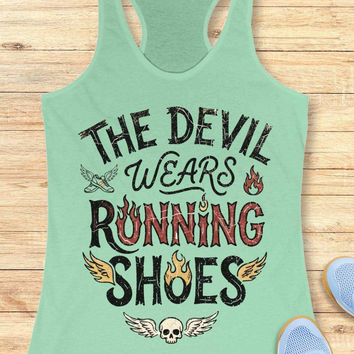 A mint green racerback tank top with a distressed graphic that reads 'THE DEVIL WEARS RUNNING SHOES' in black and red lettering, decorated with wings, flames, and a skull. The tank top is laid flat on a light wood plank background.