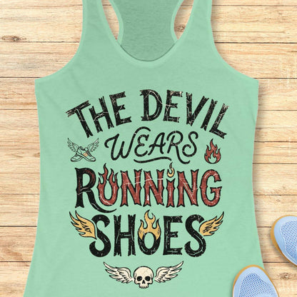 A mint green racerback tank top with a distressed graphic that reads 'THE DEVIL WEARS RUNNING SHOES' in black and red lettering, decorated with wings, flames, and a skull. The tank top is laid flat on a light wood plank background.