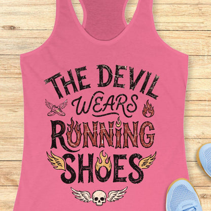 A pink racerback tank top laid flat on a light wood background, featuring the graphic text 'THE DEVIL WEARS RUNNING SHOES' in distressed black and red lettering with small flame and wing illustrations. Partial blue shoes are visible at the bottom right as styling props.