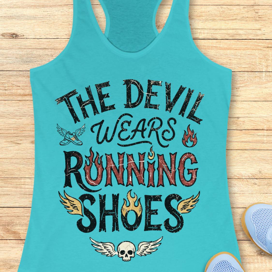 A blue racerback tank top laid flat on a light wood background, featuring the graphic text 'THE DEVIL WEARS RUNNING SHOES' in distressed black and red lettering with small flame and wing illustrations. Partial blue shoes are visible at the bottom right as styling props.