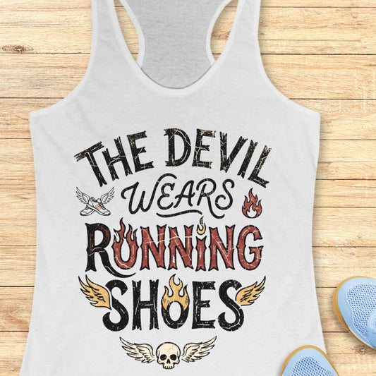A white racerback tank top laid flat on a light wood surface, printed with the phrase “THE DEVIL WEARS RUNNING SHOES” in black and red lettering with small flame and wing graphics. Part of a blue shoe is visible at the bottom right as a styling prop.
