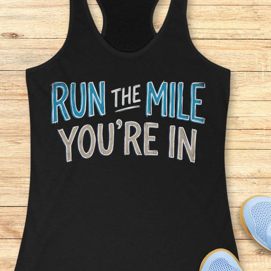 A black racerback tank top laid flat on a light wood background, featuring the motivational text 'RUN THE MILE YOU'RE IN' in orange and dark gray lettering. Partial blue sneakers are visible at the bottom right as styling props.