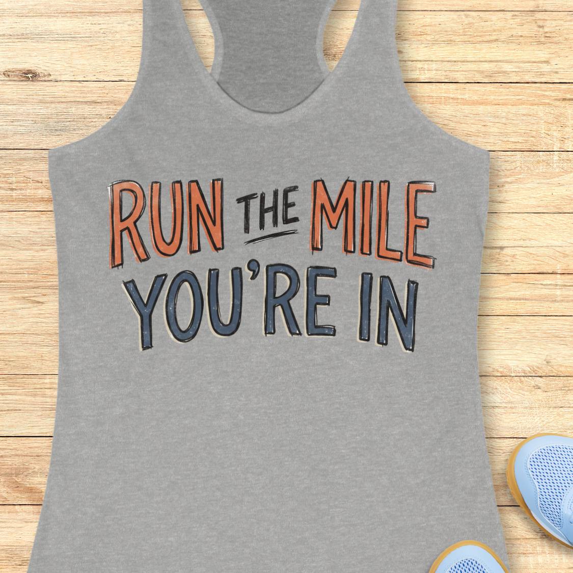 A heather gray racerback tank top laid flat on a light wood background, featuring the motivational text 'RUN THE MILE YOU'RE IN' in orange and dark gray lettering. Partial blue sneakers are visible at the bottom right as styling props.