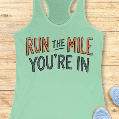 A mint racerback tank top laid flat on a light wood background, featuring the motivational text 'RUN THE MILE YOU'RE IN' in orange and dark gray lettering. Partial blue sneakers are visible at the bottom right as styling props.