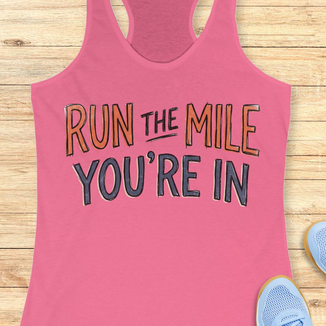 A pink racerback tank top laid flat on a light wood background, featuring the motivational text 'RUN THE MILE YOU'RE IN' in orange and dark gray lettering. Partial blue sneakers are visible at the bottom right as styling props.