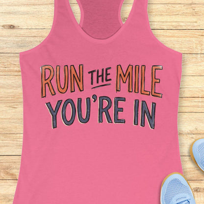 A pink racerback tank top laid flat on a light wood background, featuring the motivational text 'RUN THE MILE YOU'RE IN' in orange and dark gray lettering. Partial blue sneakers are visible at the bottom right as styling props.