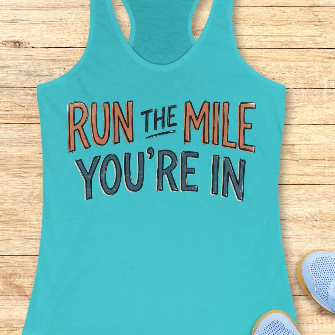 A blue racerback tank top laid flat on a light wood background, featuring the motivational text 'RUN THE MILE YOU'RE IN' in orange and dark gray lettering. Partial blue sneakers are visible at the bottom right as styling props.