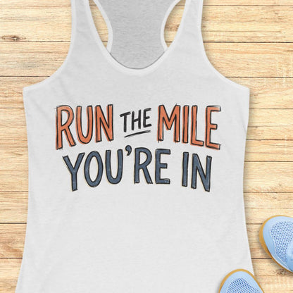 A white racerback tank top laid flat on a light wood background, featuring the motivational text 'RUN THE MILE YOU'RE IN' in orange and dark gray lettering. Partial blue sneakers are visible at the bottom right as styling props.