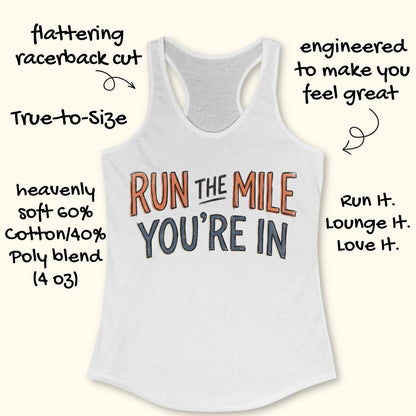 A white racerback tank top laid flat on a light wood background, featuring the motivational text 'RUN THE MILE YOU'RE IN' in orange and dark gray lettering. Partial blue sneakers are visible at the bottom right as styling props.