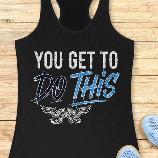 A black racerback tank top laid flat on a light wood background, featuring a graphic print that reads 'YOU GET TO DO THIS' with a pair of winged sneakers underneath.