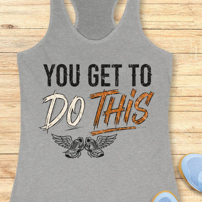 A grey racerback tank top laid flat on a light wood background, featuring a graphic print that reads 'YOU GET TO DO THIS' with a pair of winged sneakers underneath.
