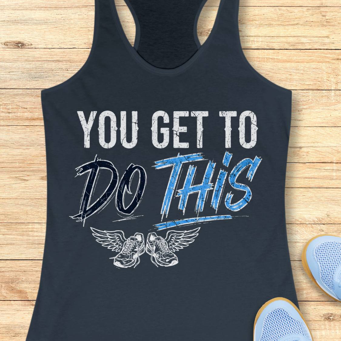 A indigo racerback tank top laid flat on a light wood background, featuring a graphic print that reads 'YOU GET TO DO THIS' with a pair of winged sneakers underneath.
