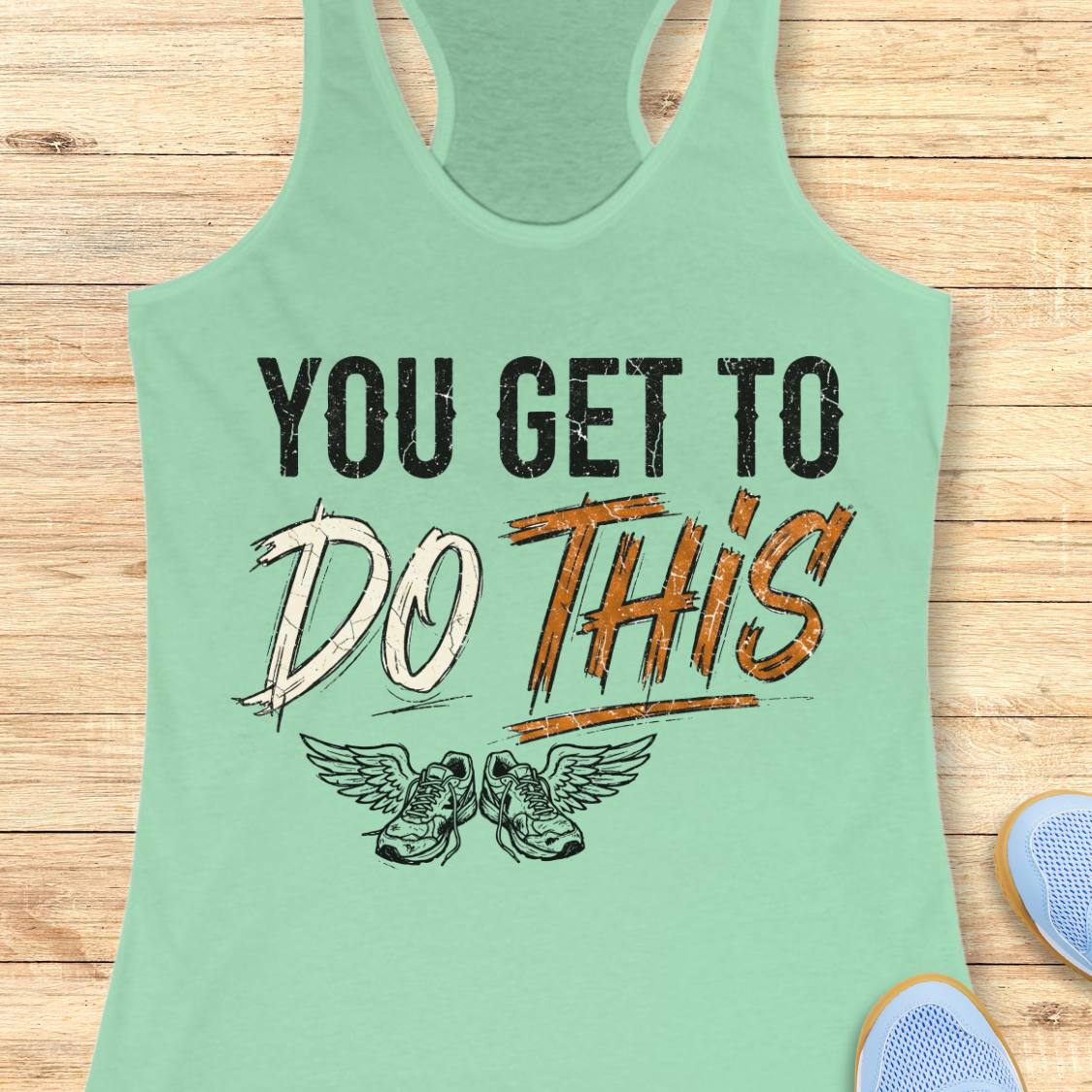 A mint green racerback tank top laid flat on a light wood background, featuring a graphic print that reads 'YOU GET TO DO THIS' with a pair of winged sneakers underneath.