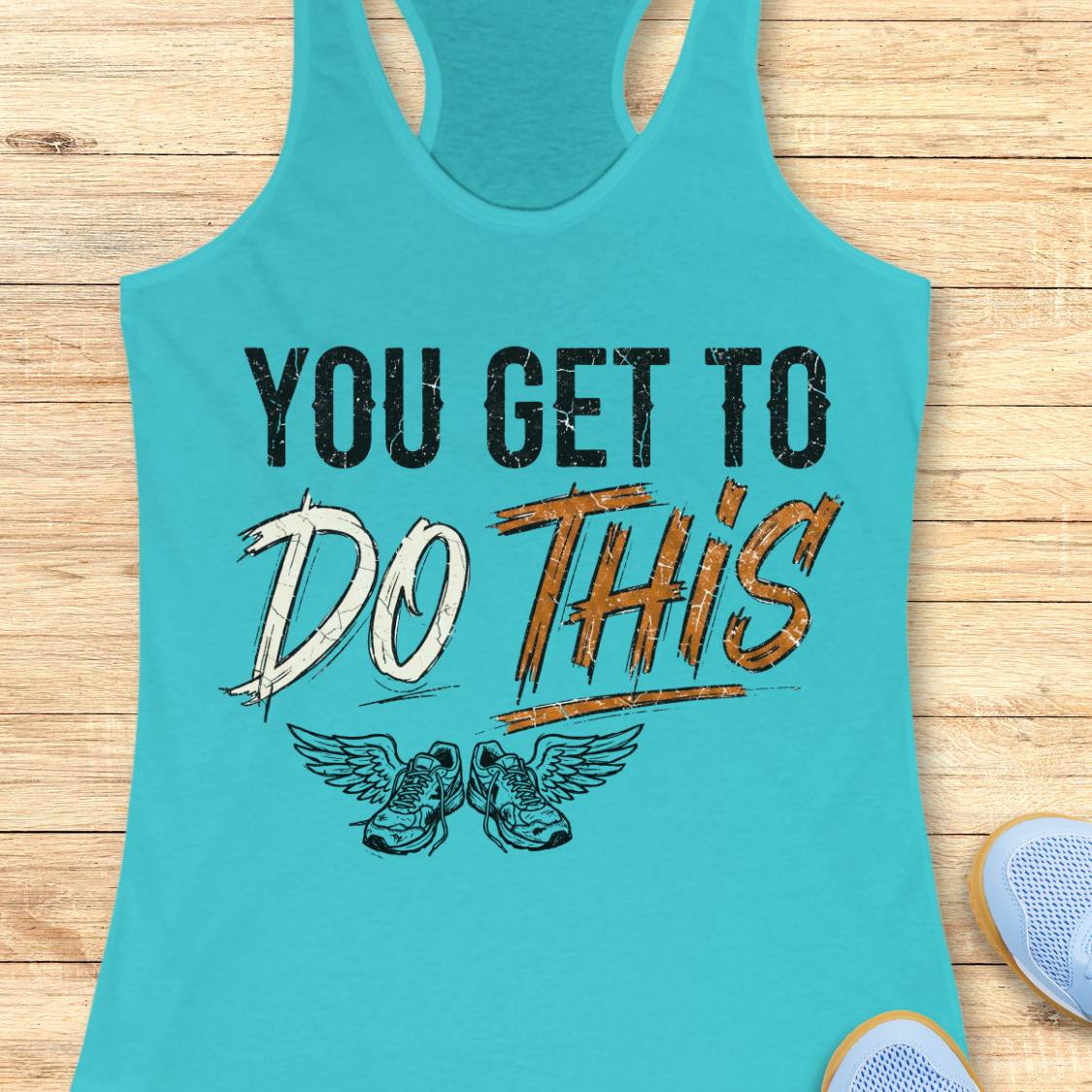 A blue racerback tank top laid flat on a light wood background, featuring a graphic print that reads 'YOU GET TO DO THIS' with a pair of winged sneakers underneath.