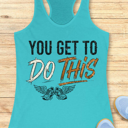 A blue racerback tank top laid flat on a light wood background, featuring a graphic print that reads 'YOU GET TO DO THIS' with a pair of winged sneakers underneath.