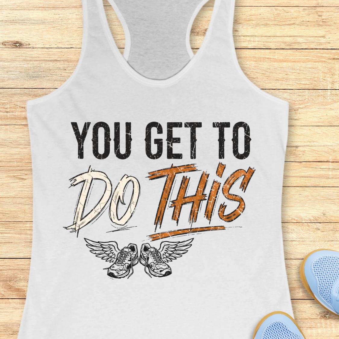 A white racerback tank top laid flat on a light wood background, featuring a graphic print that reads 'YOU GET TO DO THIS' with a pair of winged sneakers underneath.