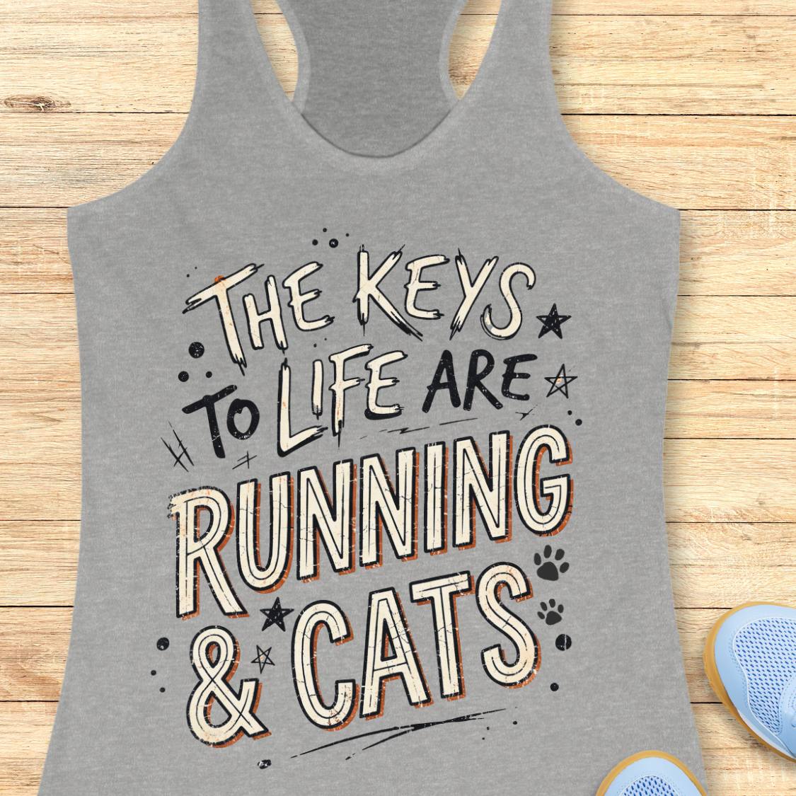 A grey racerback tank top laid flat on a light wood surface, featuring the phrase 'THE KEYS TO LIFE ARE RUNNING & CATS' in white and blue outlined lettering with small paw print and star accents. Part of a light blue shoe is visible at the bottom right.