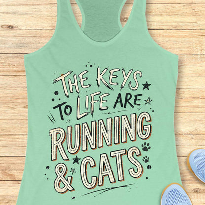 A mint racerback tank top laid flat on a light wood surface, featuring the phrase 'THE KEYS TO LIFE ARE RUNNING & CATS' in white and blue outlined lettering with small paw print and star accents. Part of a light blue shoe is visible at the bottom right.