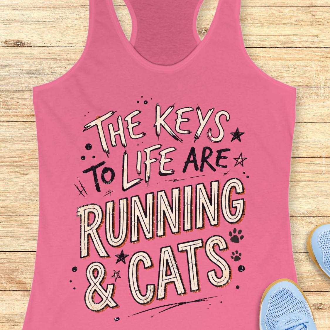 A pink racerback tank top laid flat on a light wood surface, featuring the phrase 'THE KEYS TO LIFE ARE RUNNING & CATS' in white and blue outlined lettering with small paw print and star accents. Part of a light blue shoe is visible at the bottom right.