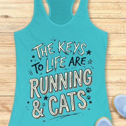 A blue racerback tank top laid flat on a light wood surface, featuring the phrase 'THE KEYS TO LIFE ARE RUNNING & CATS' in white and blue outlined lettering with small paw print and star accents. Part of a light blue shoe is visible at the bottom right.