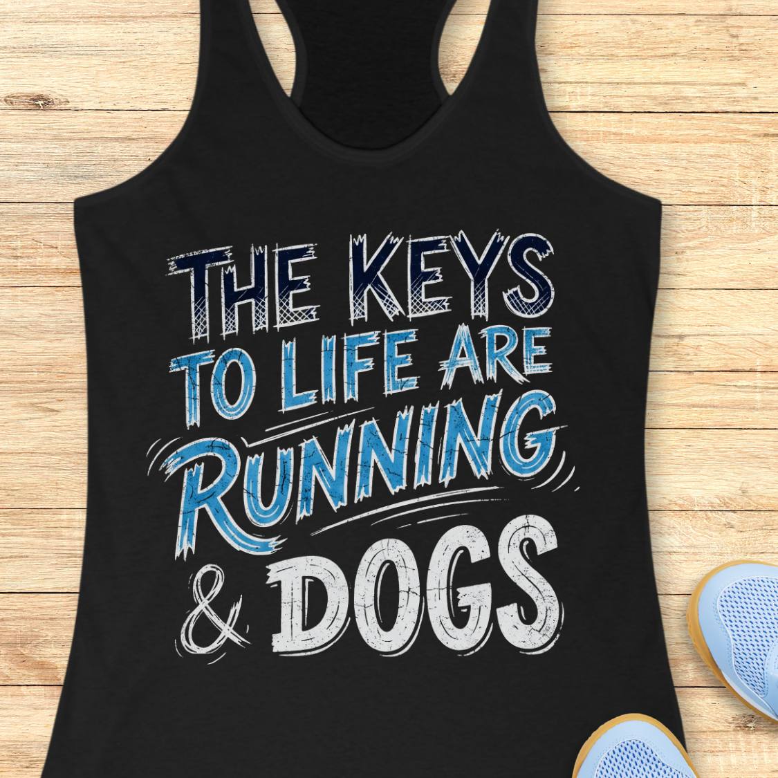 A black racerback tank top laid flat on a light wood background, featuring the text 'THE KEYS TO LIFE ARE RUNNING & DOGS' in white and blue distressed lettering. Partial blue sneakers are visible at the bottom right as styling props.