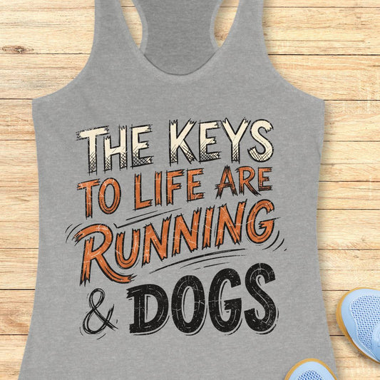 A heather gray racerback tank top laid flat on a light wood background, featuring the printed phrase “THE KEYS TO LIFE ARE RUNNING & DOGS” in white, orange, and black lettering. Partial blue shoes are visible at the bottom right as styling props.