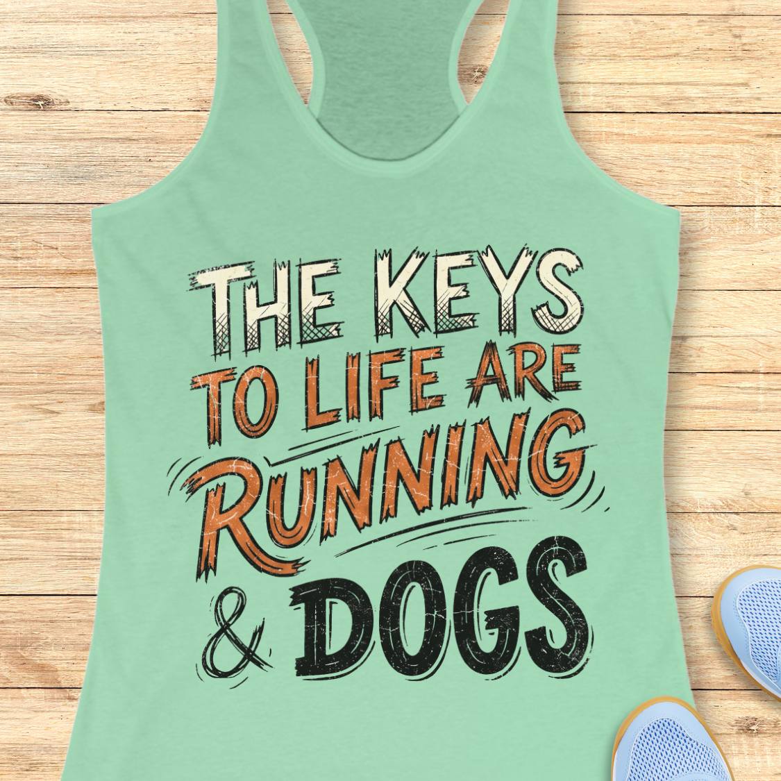 A mint racerback tank top laid flat on a light wood background, featuring the printed phrase “THE KEYS TO LIFE ARE RUNNING & DOGS” in white, orange, and black lettering. Partial blue shoes are visible at the bottom right as styling props.