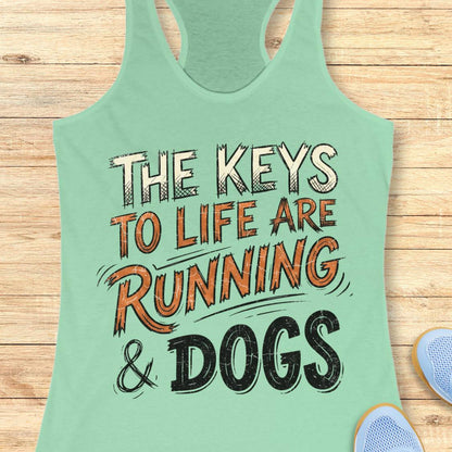 A mint racerback tank top laid flat on a light wood background, featuring the printed phrase “THE KEYS TO LIFE ARE RUNNING & DOGS” in white, orange, and black lettering. Partial blue shoes are visible at the bottom right as styling props.