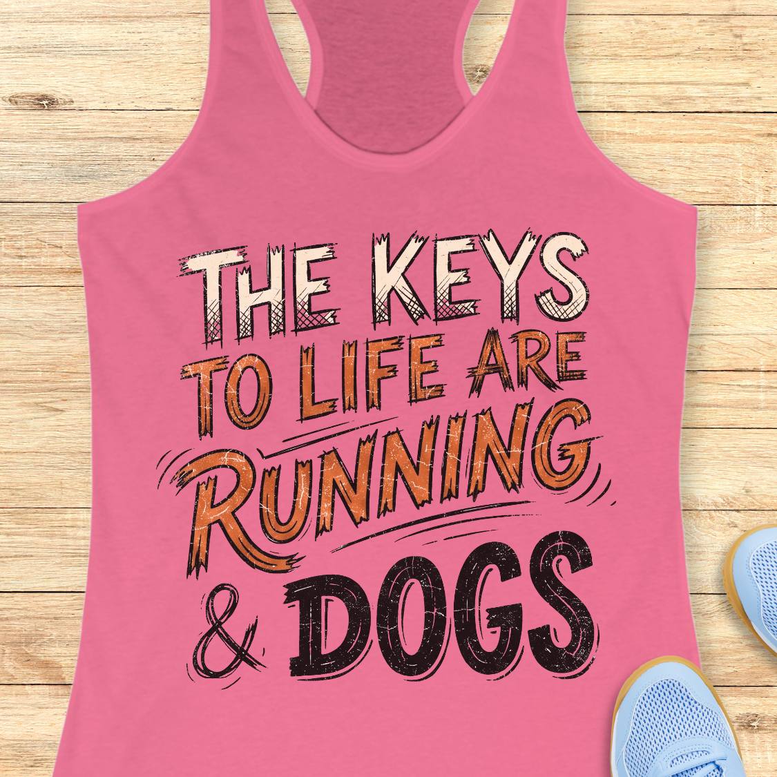 A pink racerback tank top laid flat on a light wood surface, featuring the text 'THE KEYS TO LIFE ARE RUNNING & DOGS' in a distressed graphic font. Partial blue sneakers are visible at the bottom right as styling props.
