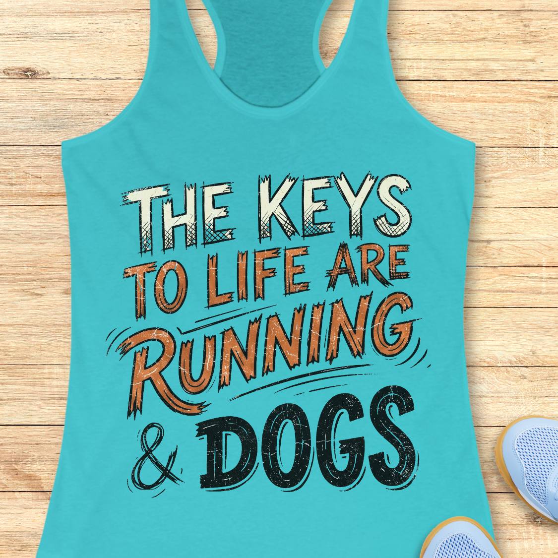 A blue racerback tank top laid flat on a light wood background, featuring the printed phrase “THE KEYS TO LIFE ARE RUNNING & DOGS” in white, orange, and black lettering. Partial blue shoes are visible at the bottom right as styling props.