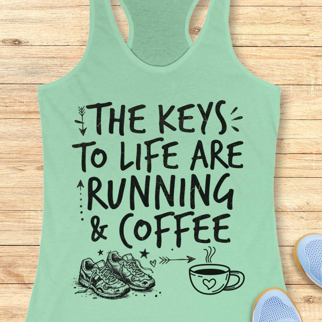 A mint green racerback tank top with a black graphic print reading 'THE KEYS TO LIFE ARE RUNNING & COFFEE' with illustrations of sneakers, stars, and a coffee cup, displayed flat on a light wood background.