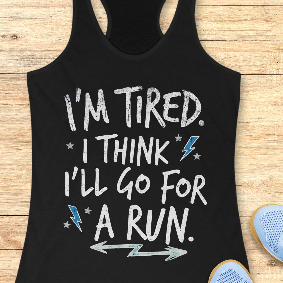 A black racerback tank top laid flat on a light wood background, with white distressed text reading “I'M TIRED. I THINK I'LL GO FOR A RUN.