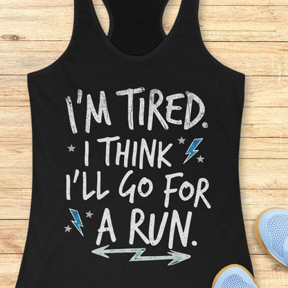 A black racerback tank top laid flat on a light wood background, with white distressed text reading “I'M TIRED. I THINK I'LL GO FOR A RUN.