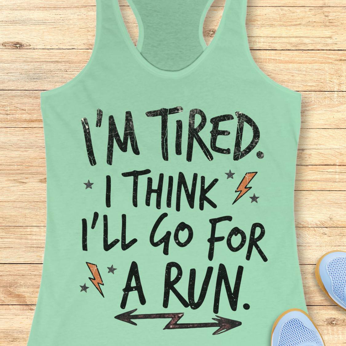 A mint green racerback tank top laid flat on a light wood background, featuring the text 'I'M TIRED. I THINK I'LL GO FOR A RUN.