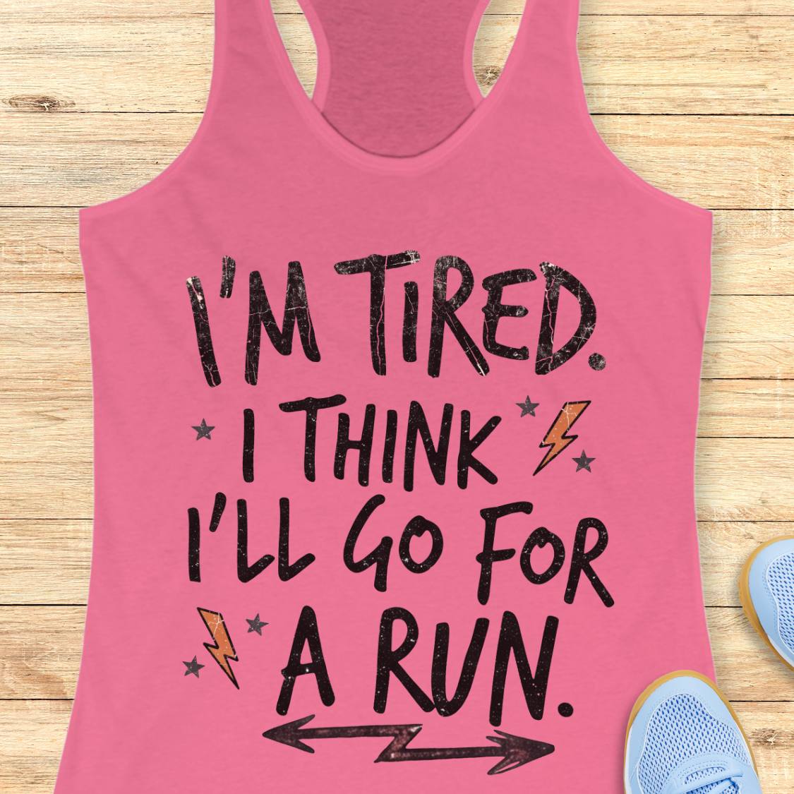 A pink racerback tank top laid flat on a light wood surface, printed with the phrase “I'M TIRED. I THINK I'LL GO FOR A RUN.