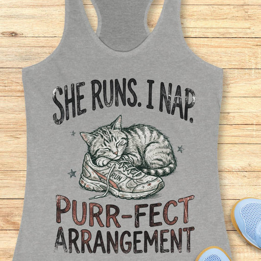 A heather gray racerback tank top laid flat on a light wood background, featuring a graphic of a sleeping cat curled up inside a sneaker with the text 'SHE RUNS. I NAP.