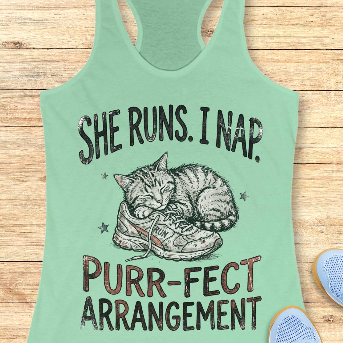 A mint green racerback tank top laid flat on a light wood background, featuring a graphic of a sleeping cat curled up inside a sneaker with the text 'SHE RUNS. I NAP.