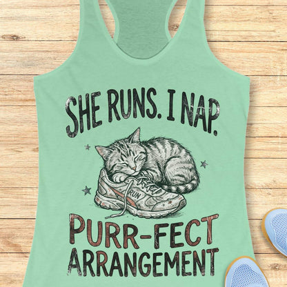 A mint green racerback tank top laid flat on a light wood background, featuring a graphic of a sleeping cat curled up inside a sneaker with the text 'SHE RUNS. I NAP.