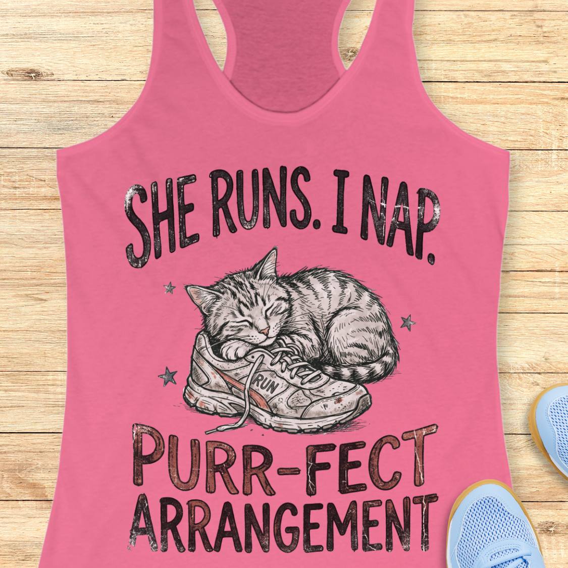 A pink racerback tank top laid flat on a light wood surface, featuring a printed graphic of a sleeping cat curled up inside a sneaker with the text 'SHE RUNS. I NAP.