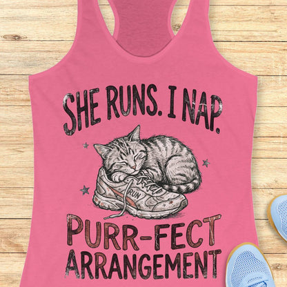 A pink racerback tank top laid flat on a light wood surface, featuring a printed graphic of a sleeping cat curled up inside a sneaker with the text 'SHE RUNS. I NAP.