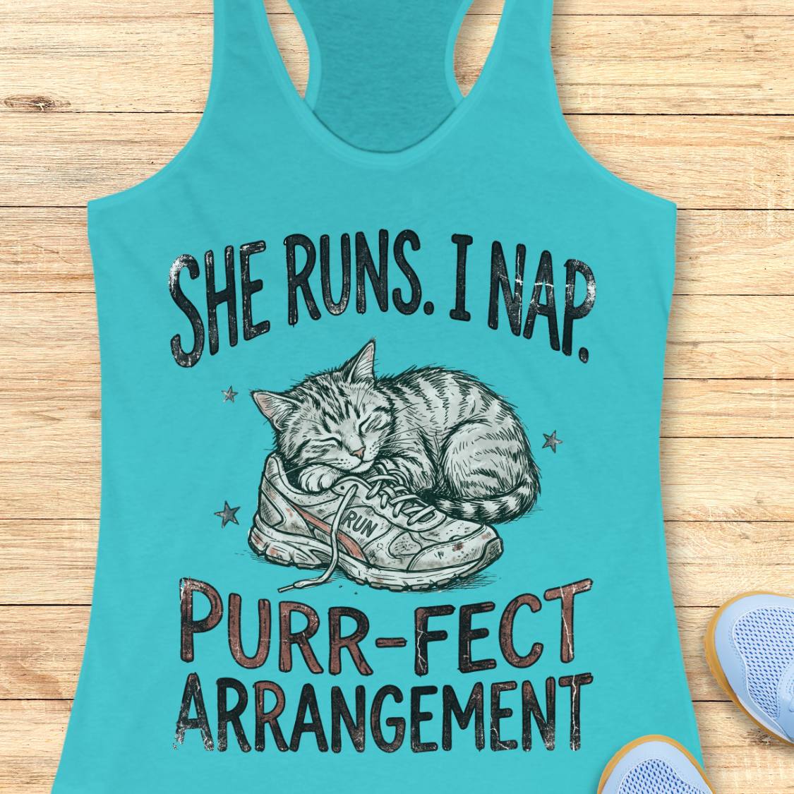 A turquoise racerback tank top laid flat on a light wood surface, featuring a graphic of a sleeping cat curled up inside a sneaker with the text 'SHE RUNS. I NAP.