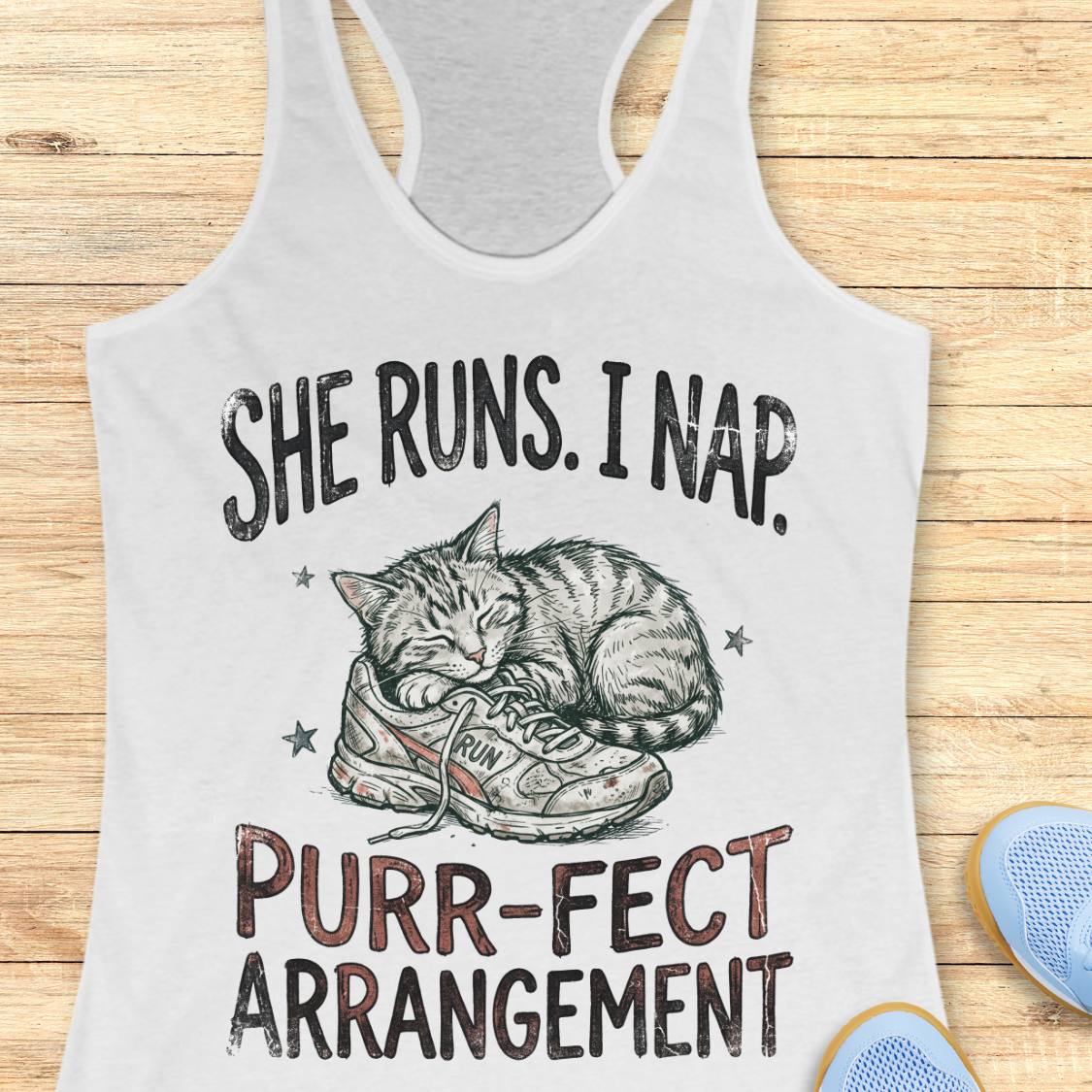 A white racerback tank top laid flat on a light wood background, featuring a printed graphic of a sleeping cat curled up around a sneaker with the text 'SHE RUNS. I NAP.