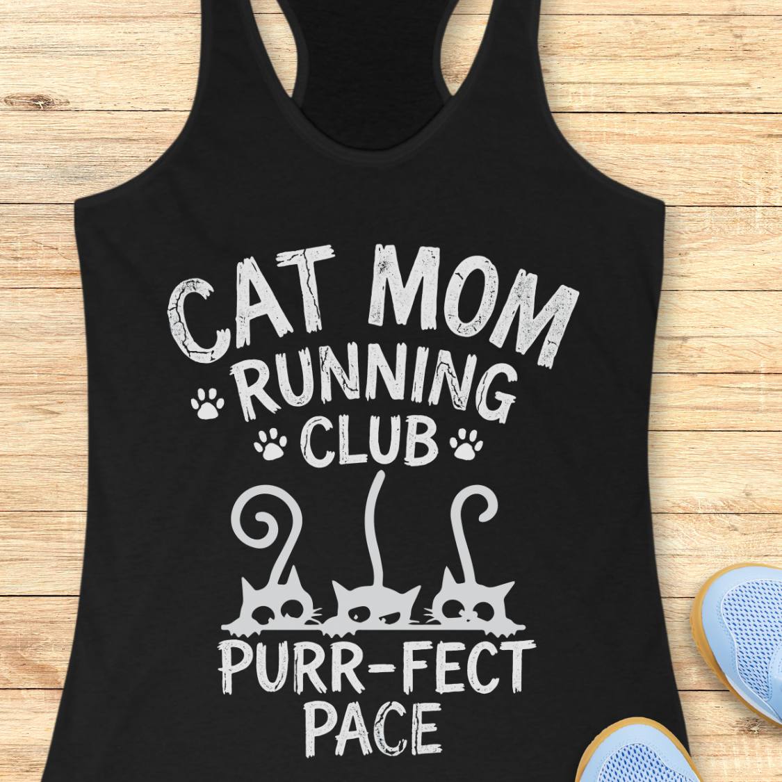 A black racerback tank top laid flat on a light wood surface, printed with the phrase 'CAT MOM RUNNING CLUB PURR-PECT PACE' in distressed white lettering, with small paw prints and cat silhouette graphics. Part of a blue shoe is visible at the bottom right.