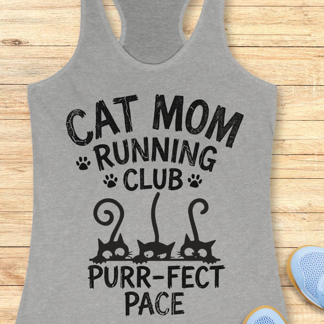 A heather gray racerback tank top laid flat on a light wood surface, printed with the phrase 'CAT MOM RUNNING CLUB PURR-FECT PACE' in black distressed lettering, with paw prints and three black cat silhouettes. Partial blue shoes are visible at the bottom right.