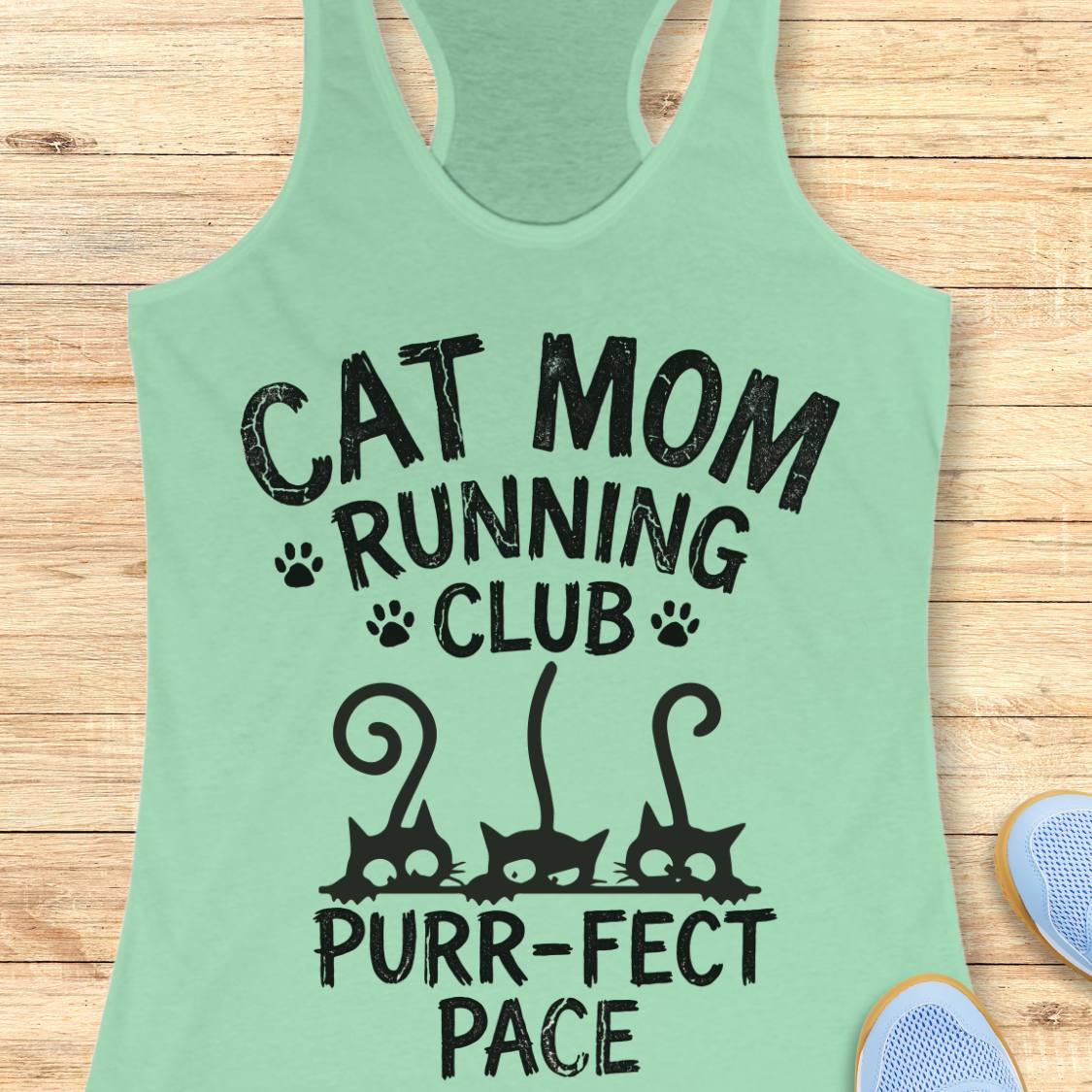 A mint green racerback tank top with a black graphic print that reads 'CAT MOM RUNNING CLUB PURR-FACT PACE' with cat silhouettes and paw prints, laid flat on a light wood background with partial shoes visible at the edges.